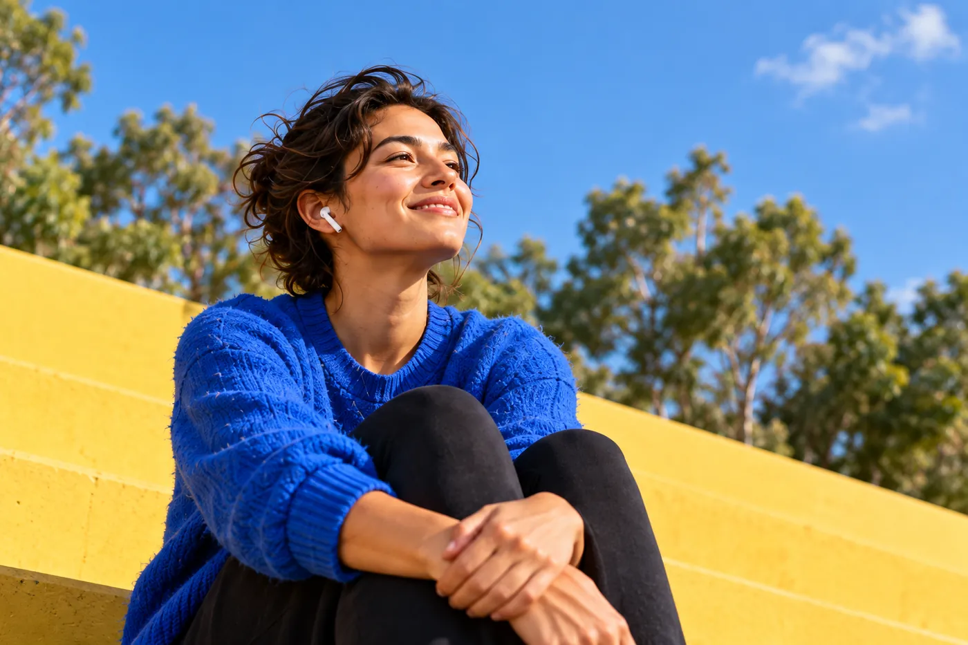 Woman with earbuds in a blue sweater sitting on yellow steps in the sun